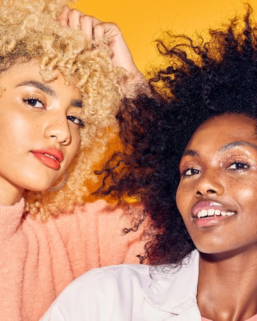 Close-up of two smiling women with natural hair against a bright yellow background; one woman has blonde curly hair, the other has dark curly hair.