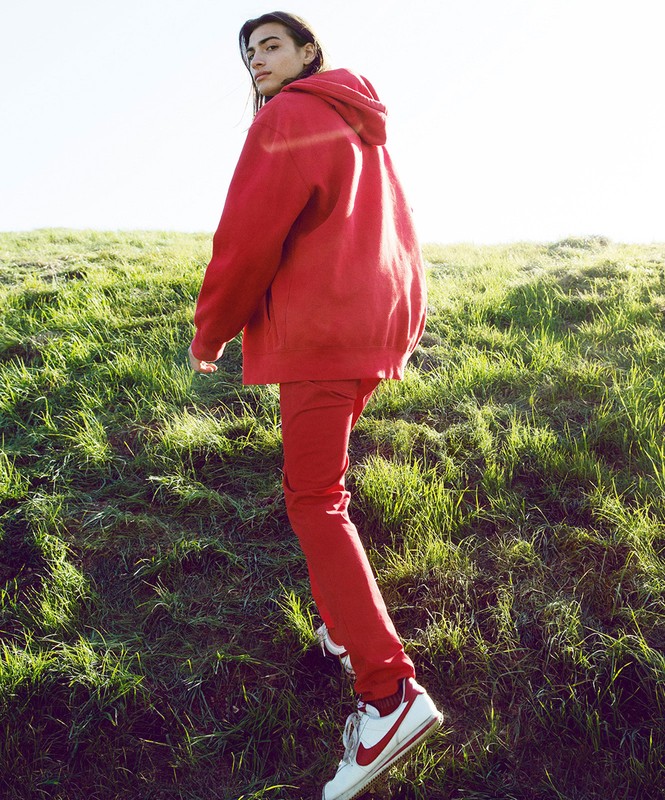 Young adult stands in grassy field wearing a red hooded sweatshirt and red pants looking over their shoulder towards the camera.