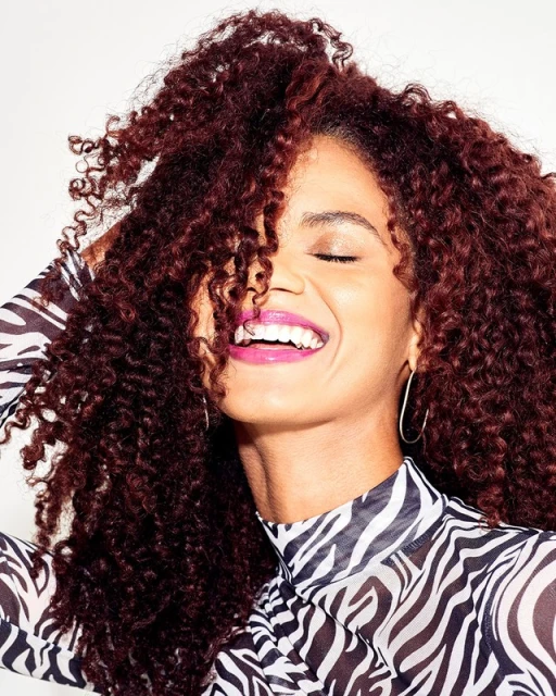 Smiling woman with curly auburn hair, wearing a zebra-striped shirt, posing against a white backdrop in a beauty-style headshot.