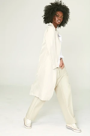 Full-length studio shot of a Black female model in a long cream-colored coat, cream wide-leg pants and white sneakers. She is walking forward.