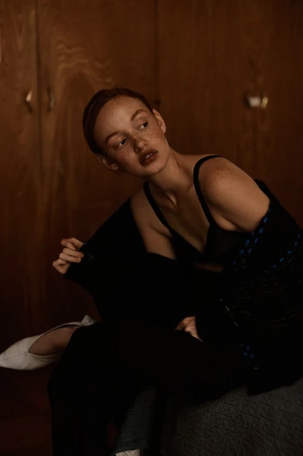 Woman with freckles poses in a black bra and cardigan in an indoor setting with wood paneling, exuding natural beauty and confidence.