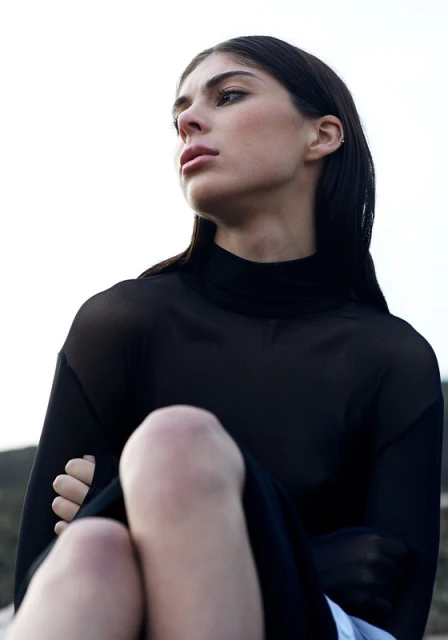Young woman with long dark hair, wearing a black mesh top, poses with her arms crossed and knees up against a bright sky backdrop.