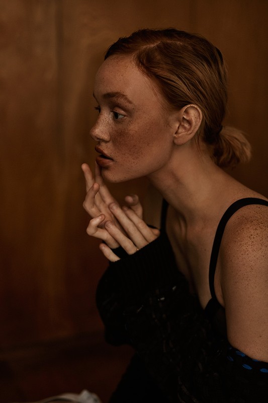 Profile portrait of a light-skinned woman with freckles, fair hair in a low bun, and a black dress, in an indoor setting with warm lighting.