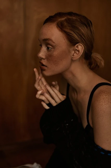 Profile portrait of a light-skinned woman with freckles, fair hair in a low bun, and a black dress, in an indoor setting with warm lighting.