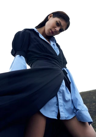 Low angle portrait of a woman wearing a black wrap dress over a light blue buttoned shirt against a sky background