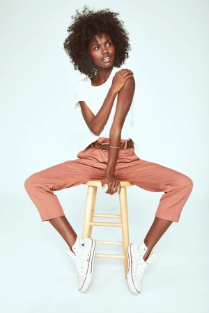 Black woman with voluminous afro hair wearing a white t-shirt and pink trousers, seated on a wooden stool.