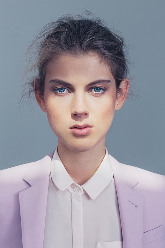 Portrait of a fair-skinned young woman with blue eyes wearing a lavender blazer and white collared shirt against a grey background.