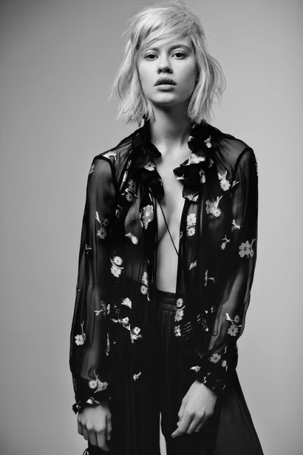 Black-and-white portrait of a young adult woman with tousled blonde hair, wearing a sheer floral blouse, posing in a photo studio.