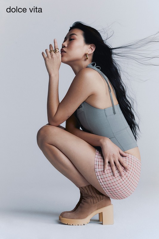 A young Asian woman crouches indoors wearing a grey top, a plaid skirt, and platform boots, showcasing fashion and style in a studio setting.