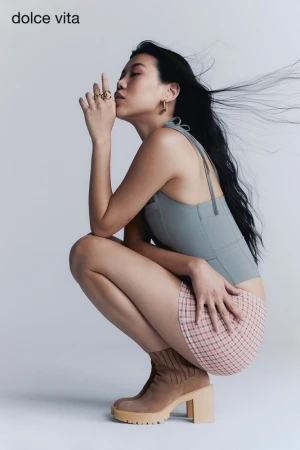 Model squatting in a grey halter top and plaid skirt with brown heeled boots against a white studio background