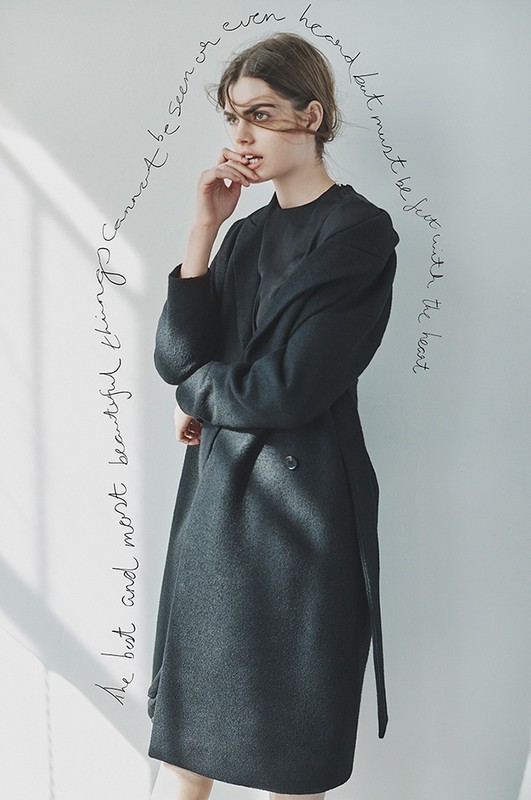 Adult female in a long black wool coat poses indoors near a window with a hand to her mouth. Soft daylight, neutral background, fashion style.