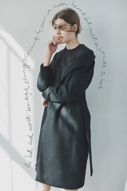 Adult female in a long black wool coat poses indoors near a window with a hand to her mouth. Soft daylight, neutral background, fashion style.