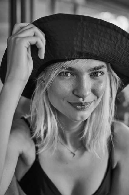 Monochrome portrait of a woman with blonde hair, wearing a dark hat and a spaghetti-strap top, looking at the camera with a slight smile.