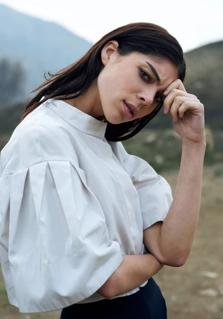 Woman in a relaxed pose outdoors, wearing a short-sleeved blouse, with mountains blurred in the background under an overcast sky.