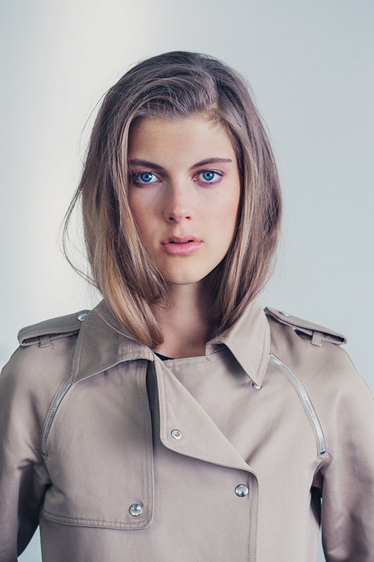 Close-up portrait of a young adult woman with blue eyes and blonde hair, wearing a tan trench coat against a neutral background.
