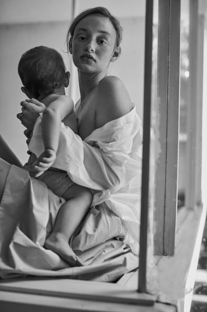 Black and white portrait of a woman holding a baby, both partially covered in a white cloth, seated near a window with natural lighting.