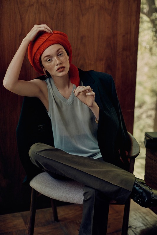 Portrait of a Caucasian woman wearing a red headscarf and a grey tank top, seated in a chair with a dark jacket draped over it.