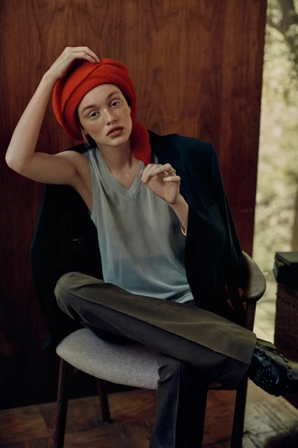 Portrait of a Caucasian woman wearing a red headscarf and a grey tank top, seated in a chair with a dark jacket draped over it.
