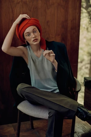 Portrait of a Caucasian woman wearing a red headscarf and a grey tank top, seated in a chair with a dark jacket draped over it.