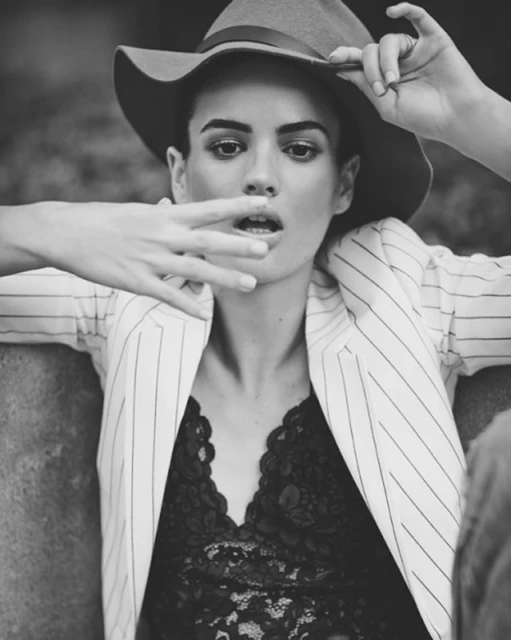 Black and white portrait of a model wearing a hat, pinstripe jacket, and lace top, with her hand near her mouth and a blurred background.