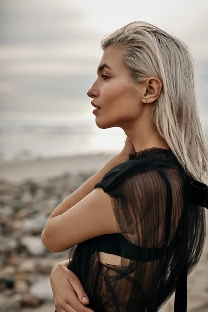Side profile of a woman with blonde hair standing on a rocky beach near ocean water, wearing a black dress against an overcast sky.