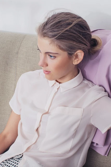 Woman in a light pink blouse seated on a beige sofa, gazing away in a studio setting with a light purple garment nearby.