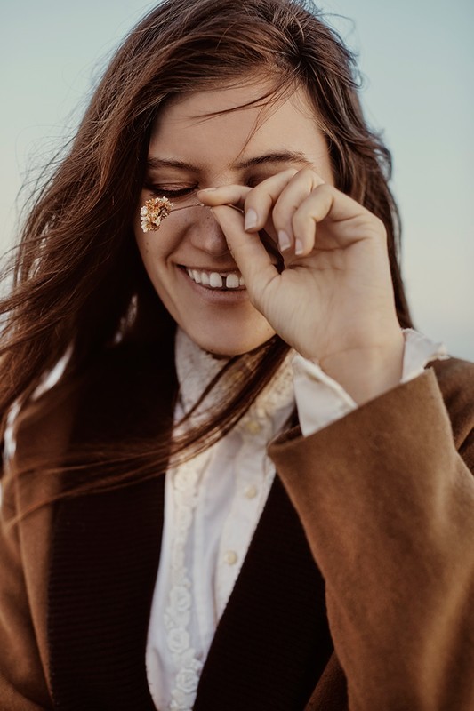 A young woman smiles outdoors, wearing a brown coat, holding a dried flower near her eye, with a blurred, light blue sky in the background.