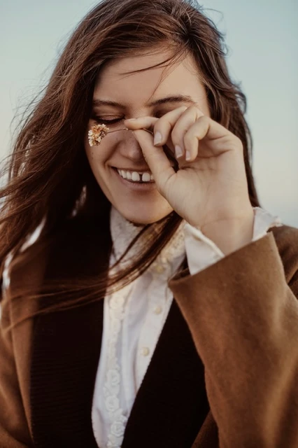 A young woman smiles outdoors, wearing a brown coat, holding a dried flower near her eye, with a blurred, light blue sky in the background.