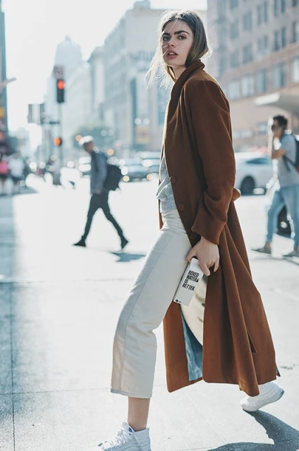 A woman with a long brown coat walks across a city street. She holds a white water bottle, and wears white shoes and cream pants.