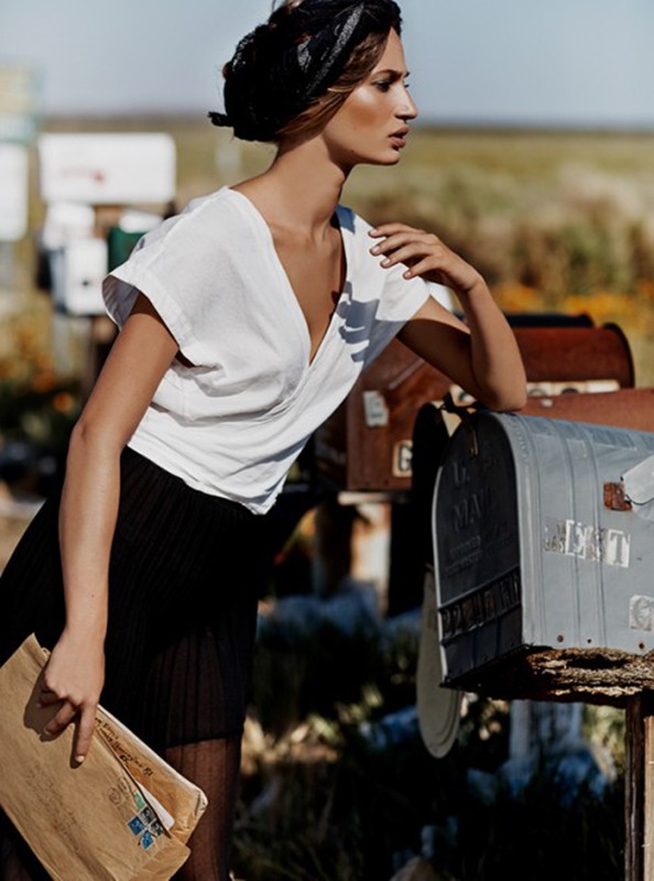 Woman in a white top and black skirt stands next to mailboxes in an outdoor setting, holding an envelope, creating an editorial style image.