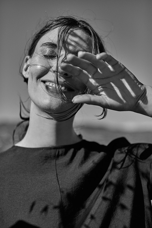 Monochrome portrait of smiling woman with hand shielding her face from sunlight, casting shadows, outdoors against clear sky background.