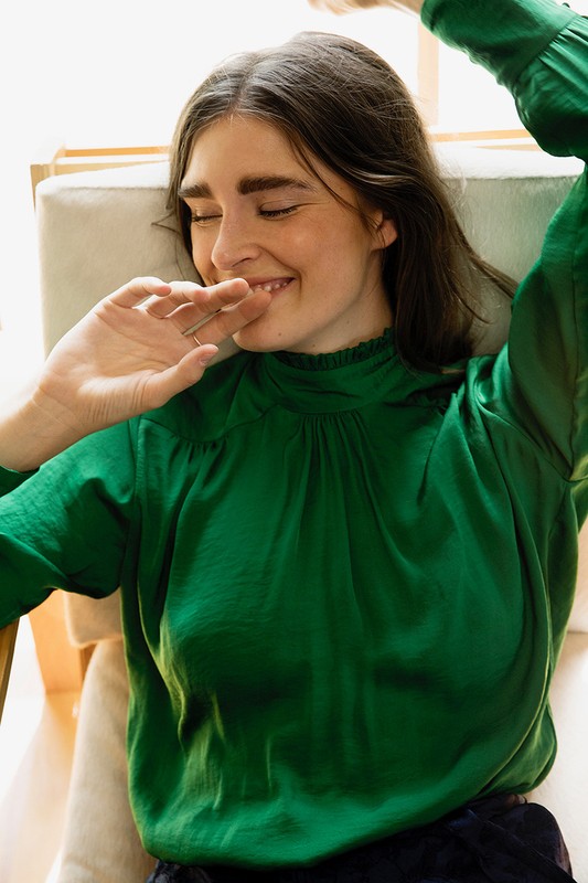 Adult woman in green long-sleeve top reclines in cushioned chair. Smiling woman with hand near face looks to the side.