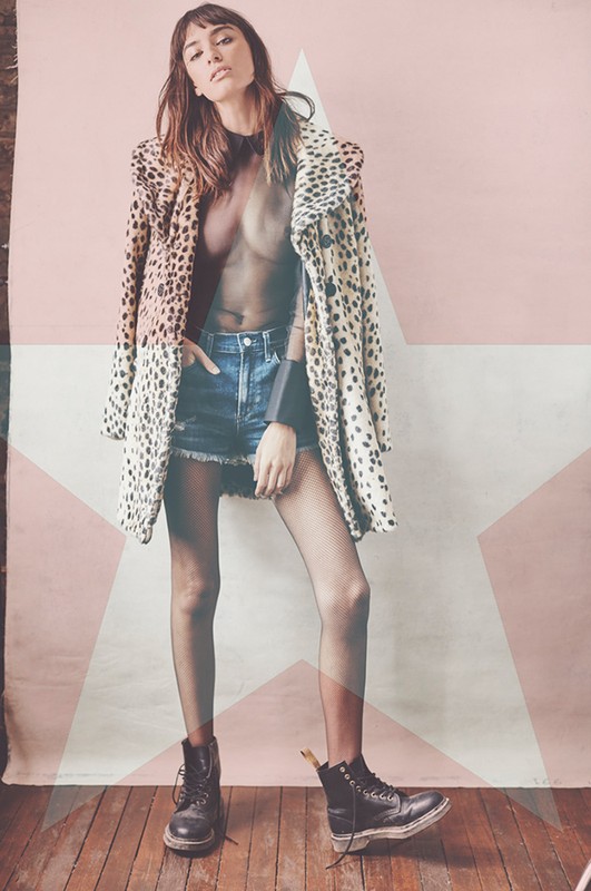 A woman in a leopard coat, black top, and denim shorts poses in front of a pink star background, wearing boots, striking a confident pose.