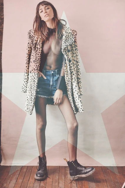 A woman in a leopard coat, black top, and denim shorts poses in front of a pink star background, wearing boots, striking a confident pose.