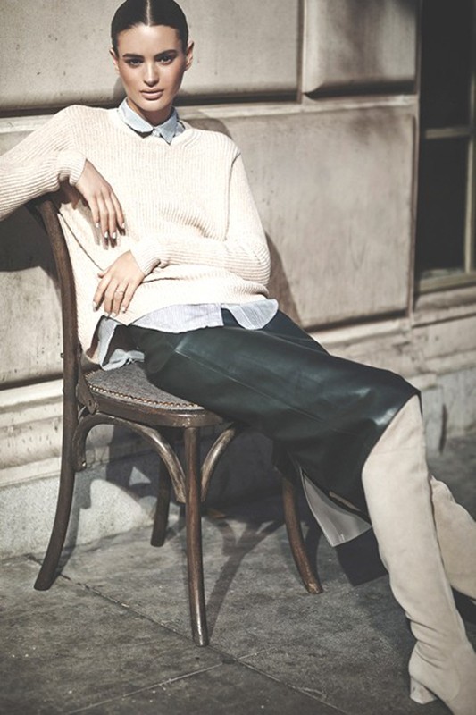 A stylish woman in an off-white sweater, black skirt, and tall beige boots sits on a wooden chair against a neutral wall.