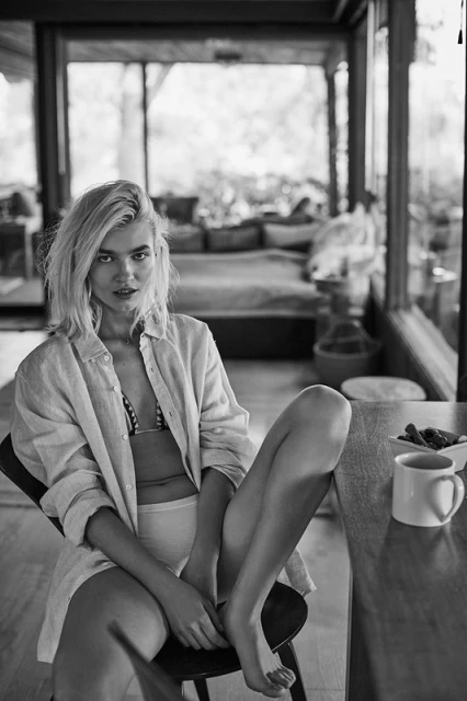 Black and white portrait of a blonde woman in a shirt and bikini top sitting on a chair inside a sunroom next to a table.