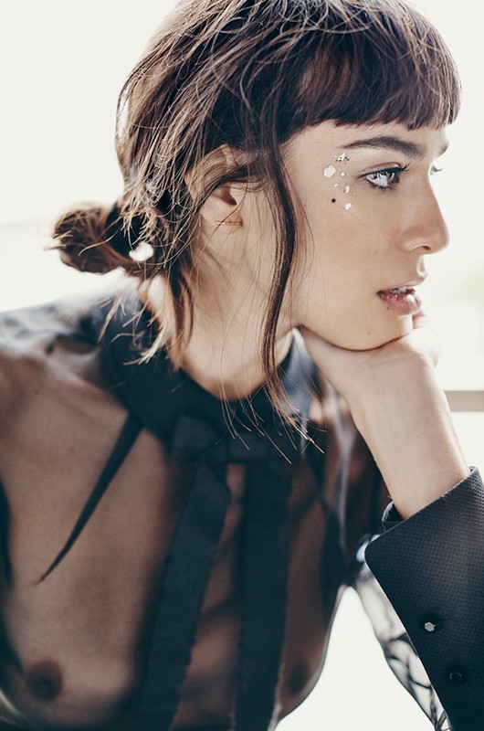 Portrait of a woman with glitter makeup wearing a sheer black top and her hair in a low bun against a blurred out background.