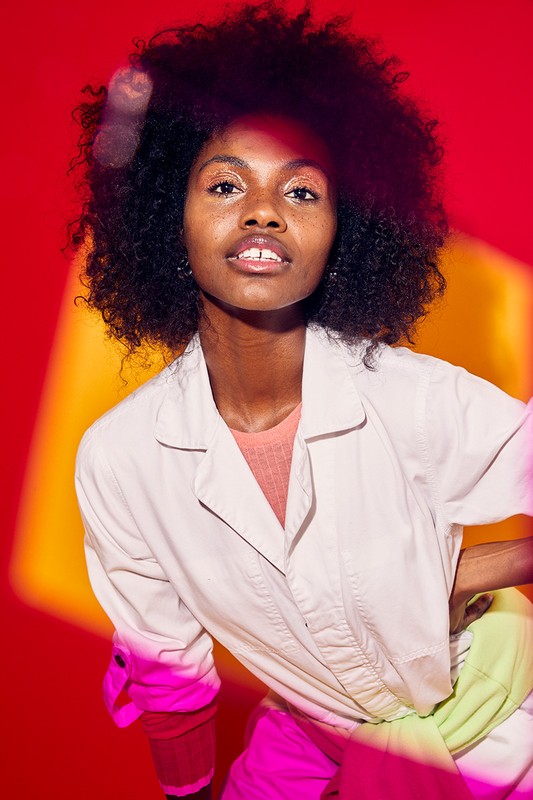 Close-up portrait of a black woman with an afro hairstyle, wearing a lab coat, in a brightly lit studio with a vibrant red backdrop.