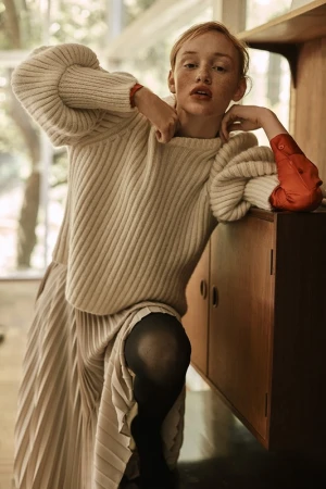 A stylish woman poses indoors wearing a cream-colored sweater, pleated skirt, and black tights, with a vintage cabinet behind her.
