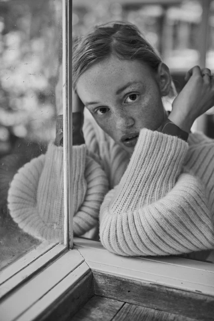 Black and white portrait of a freckled young woman wearing a sweater, looking directly at the camera while resting on a window sill.