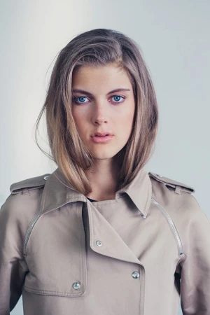 Portrait of a young woman with blue eyes wearing a beige trench coat against a neutral light background