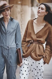 Two women posing outdoors, one wearing a grey jumpsuit and hat, the other in a brown cardigan and polka dot trousers