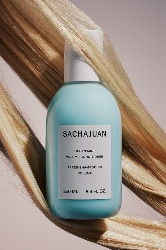 Sachajuan Ocean Mist Volume Conditioner in a blue bottle with blond strands of hair in the background on a pale pink background.