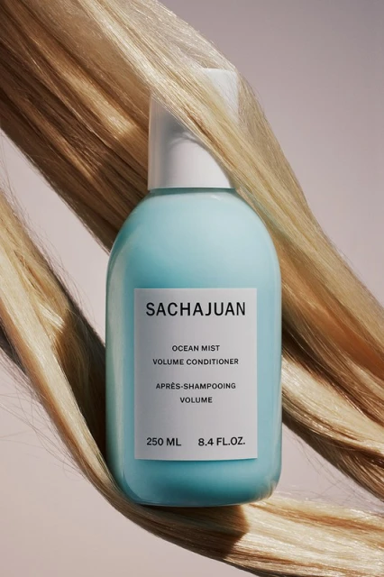 Sachajuan Ocean Mist Volume Conditioner in a blue bottle with blond strands of hair in the background on a pale pink background.