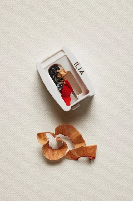 Close-up of an Ilia beauty pencil sharpener with shavings, set against a textured cream background in a simple studio shot.