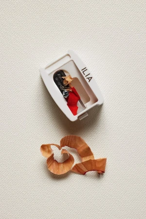 Close-up of an Ilia beauty pencil sharpener with shavings, set against a textured cream background in a simple studio shot.