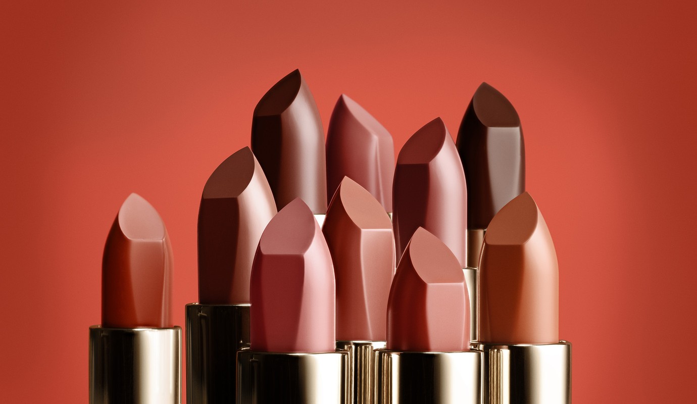 Elevated studio shot of multiple lipsticks with golden tubes in varying shades of brown and red against a solid reddish-brown backdrop.
