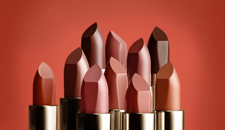 An arrangement of ten various nude and brown lipsticks in gold tubes against a solid terracotta orange background.