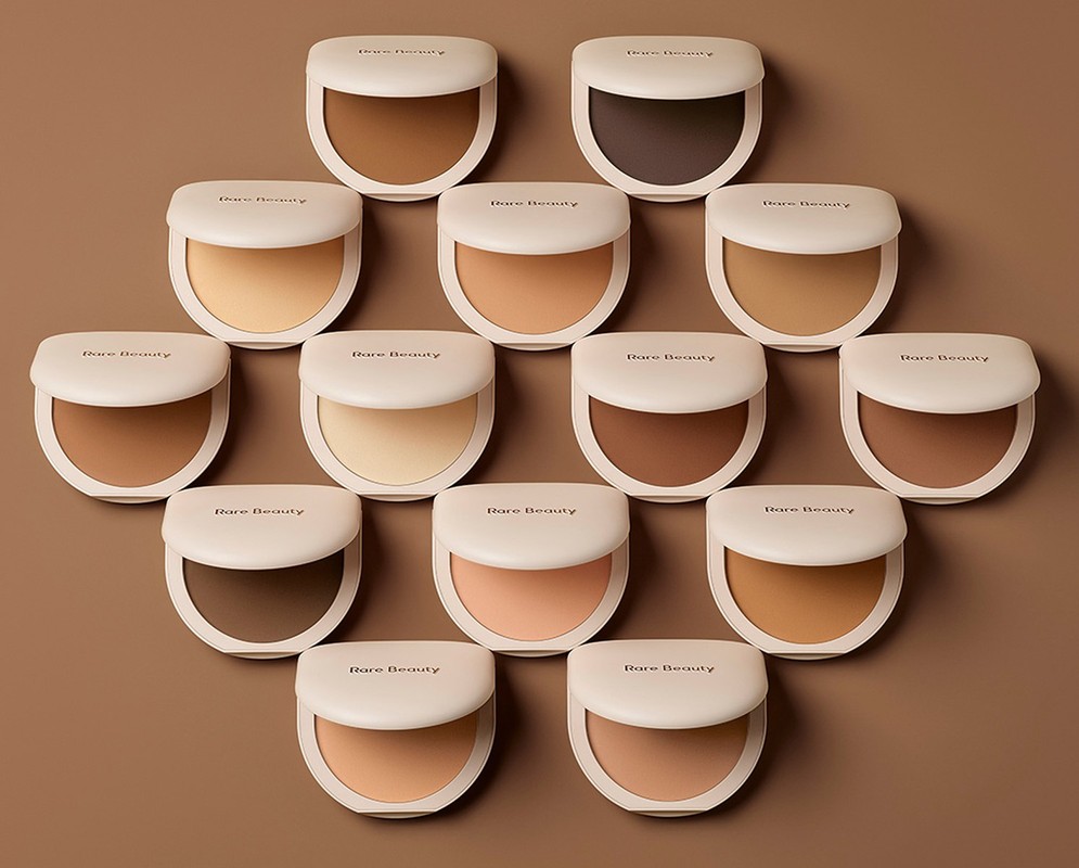 A collection of Rare Beauty compacts arranged on a neutral background, showcasing a range of foundation shades for diverse skin tones.