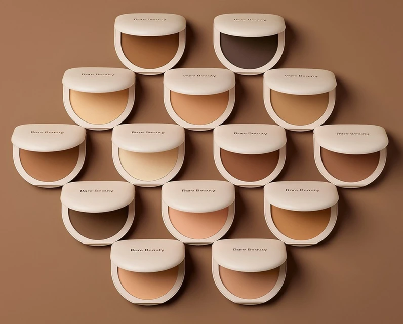 A collection of Rare Beauty compacts arranged on a neutral background, showcasing a range of foundation shades for diverse skin tones.